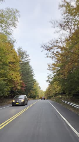 Point of view drive along a scenic mountain highway during peak autumn foliage, with colorful fall trees lining the road just before sunset as traffic moves steadily through the landscape.
