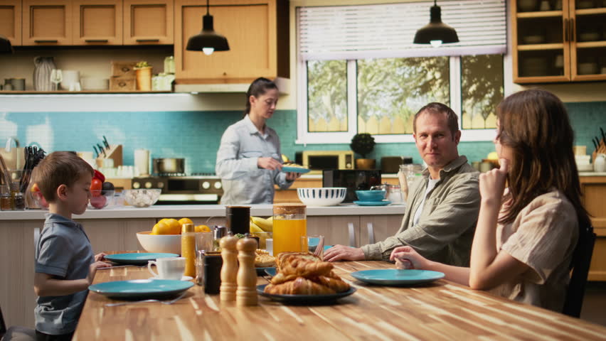 Family sharing scrambled eggs and pastry for breakfast at home, parents serving omelettes and toast with little boy and pre-teen girl. Warmth, relaxation and joyful togetherness. Camera A.