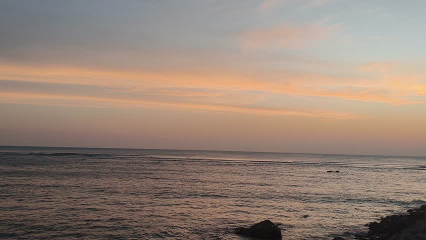 Cinematic scenic sunset over the ocean with pink and orange clouds. Calm sea waves flowing near silhouette rocks during golden hour twilight. Peaceful and dramatic nature background.