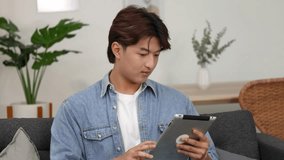 A young Asian man in a denim shirt uses a tablet while sitting indoors - Powered by Shutterstock - Get 15% off with code: PIKWIZARD15