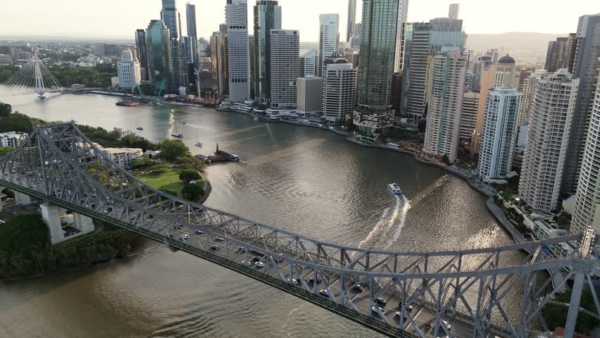 A wide drone shot looking across the Brisbane River toward the central business district. The Story Bridge is in the foreground with traffic moving across it, while a river ferry cruises past high-rise skyscrapers during a golden sunset.