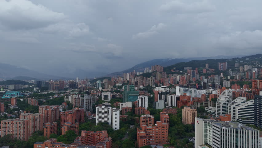 Drone shot capturing the striking Medellin El Poblado skyline against moody clouds. High-rise buildings merge with lush greenery, offering a vibrant urban landscape scene in Colombia.