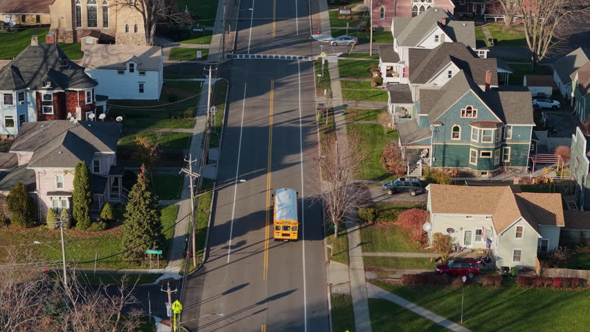 Wilson, NY, USA, 20 October 2025: Aerial view of yellow school bus driving through quiet American residential neighborhood with houses, church buildings, crosswalk and autumn trees, everyday suburban
