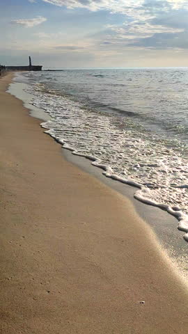 Seascape on sunny summer day. Sea waves rolling on sand of sandy beach on seashore. Waves of quiet calm sea, sand with footprints, blue sky with clouds and horizon line. Vertical