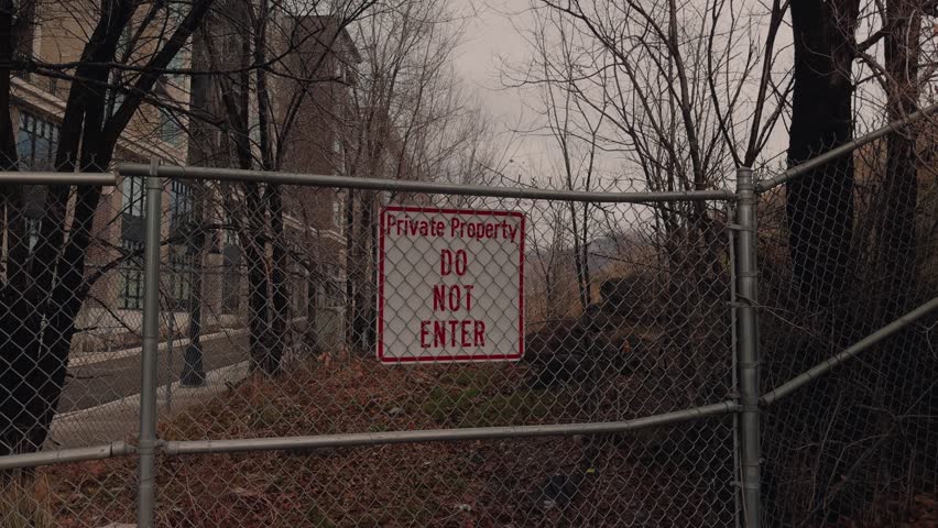 Slow panning shot of a private property warning sign on a chain link fence near trees.