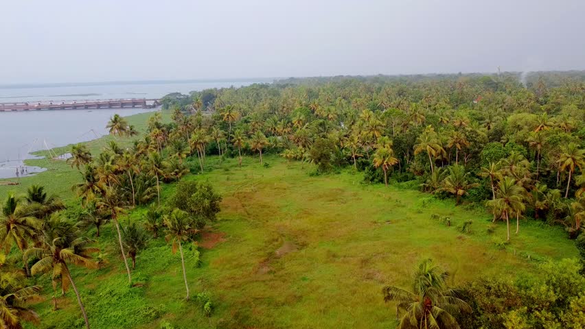 Aerial view of lush green Kerala landscape with coconut palms, open fields, and a distant bridge over backwaters.