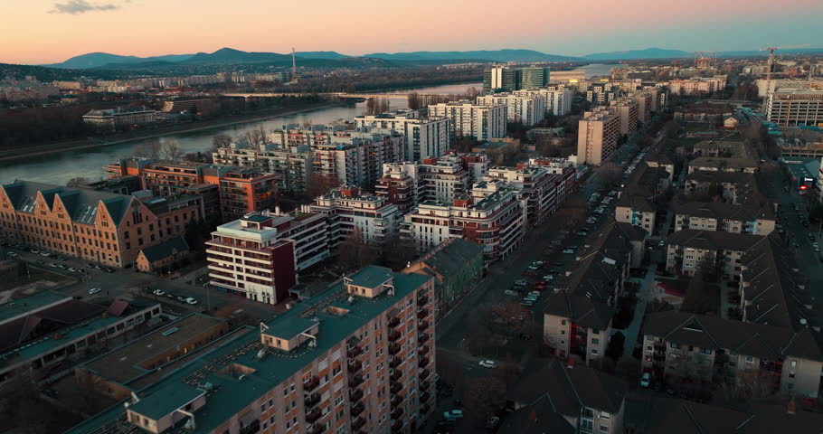 Aerial shot of Budapest showing residential apartment blocks with the Danube River in the background and hills on the horizon. Urban housing, city layout, and natural landscape visible on a clear day.