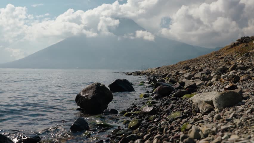 Rocky Lake Atitlan shoreline with gentle waves and San Pedro Volcano under clouds