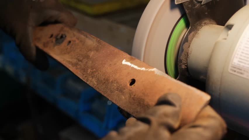 A detailed slow-motion close-up shows a steel lawn mower blade being sharpened on a bench grinder. These intense sparks are ideal for hardware commercials, tool maintenance guides, and industrial ads.
