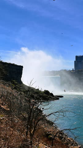 View of Niagara Falls with mist rising and birds flying around near the water on a sunny day showcasing nature power and beauty.