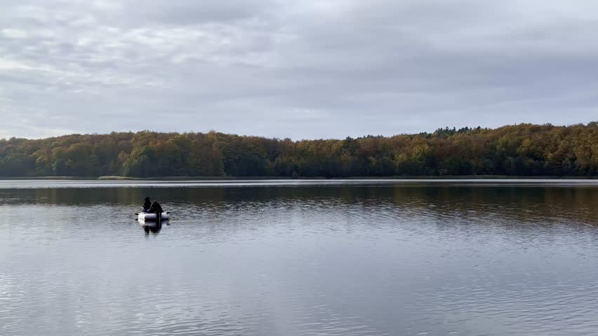 Two unrecognizable fishermen are sitting on a small inflatable boat while fishing on a calm lake in Poland. The wide shot shows still water with soft reflections, a forested shoreline in autumn colors, and an overcast sky. The scene conveys quiet leisure, outdoor recreation, and peaceful freshwater fishing in a natural landscape. Suitable for concepts related to fishing, relaxation, rural life, and nature-based activities.
