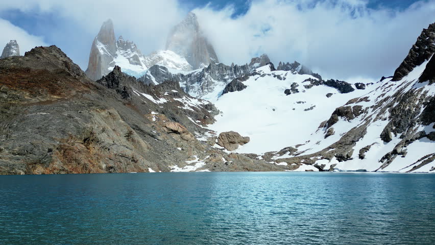 Aerial drone view of a turquoise mountain lake surrounded by rugged terrain and snowfields beneath the Fitz Roy massif