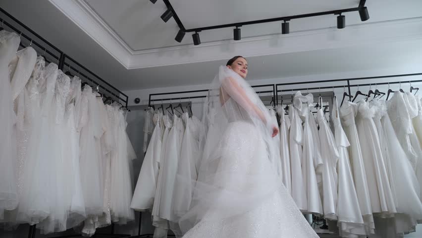 A luxurious female model in a wedding dress with beautiful makeup poses with a long veil in a modern wedding salon, she spins and looks at the camera. Stabilized image from below.