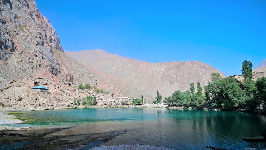 A view of Khurdak Lake, the fifth lake of the Seven Lakes in the Fann Mountains near Panjakent, Tajikistan, with blue water, rocky slopes, and narrow mountain terrain under clear daylight.