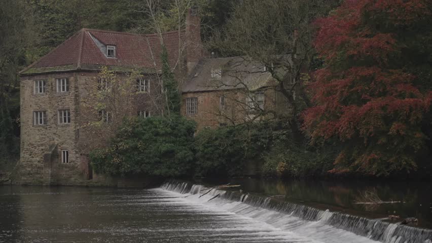 A static medium shot focusing on the historic stone Fulling Mill on the banks of the River Wear, with water cascading over the famous weir in the foreground on a moody autumn morning in Durham.