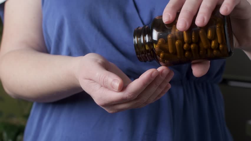Woman pours dietary supplement capsules from amber bottle into open palm indoors, illustrating wellness nutrition concept.