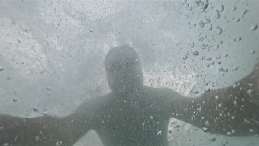 Submerged man wearing swim goggles moves underwater among bubbles as waves pass above during sea diving with a conceptual drowning theme.
