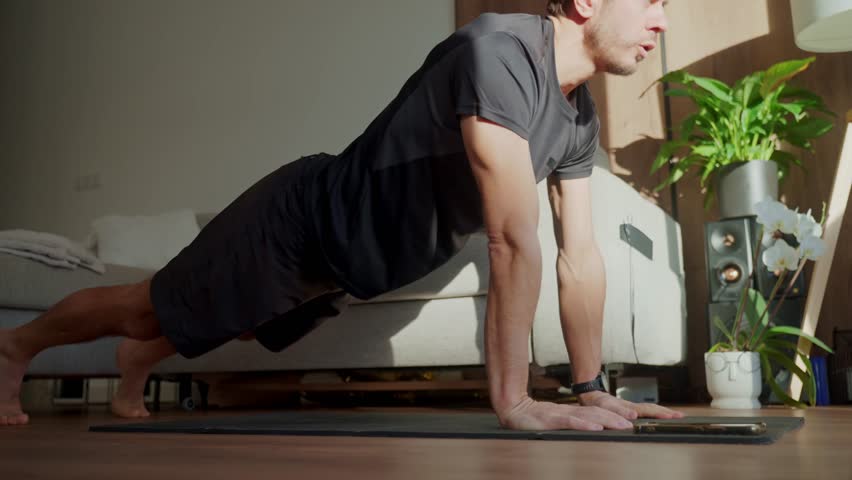 Man in athletic wear doing up down plank exercise on fitness mat in living room. Concept of home workout, cardio training, bodyweight exercise and healthy lifestyle