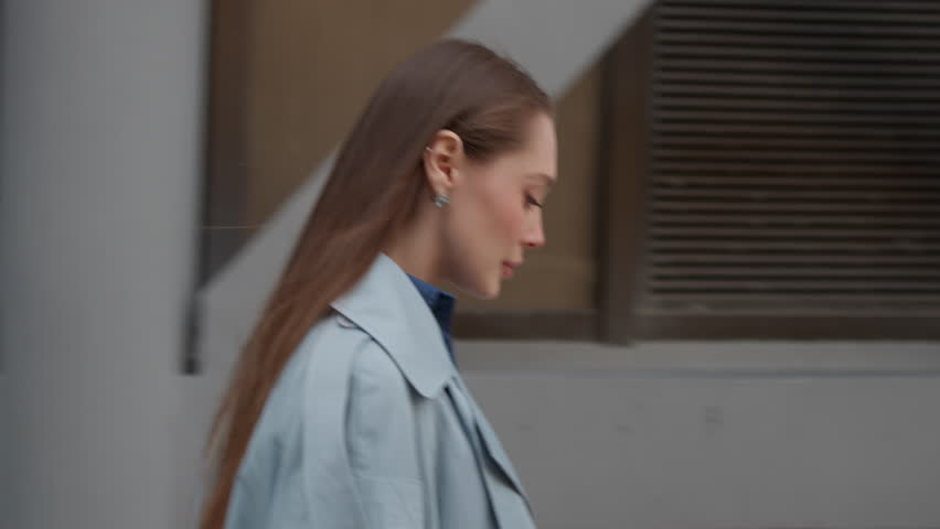 Side view of young woman with long hair walking on city sidewalk in stylish blue coat. Beautiful lady is moving through urban street during daytime