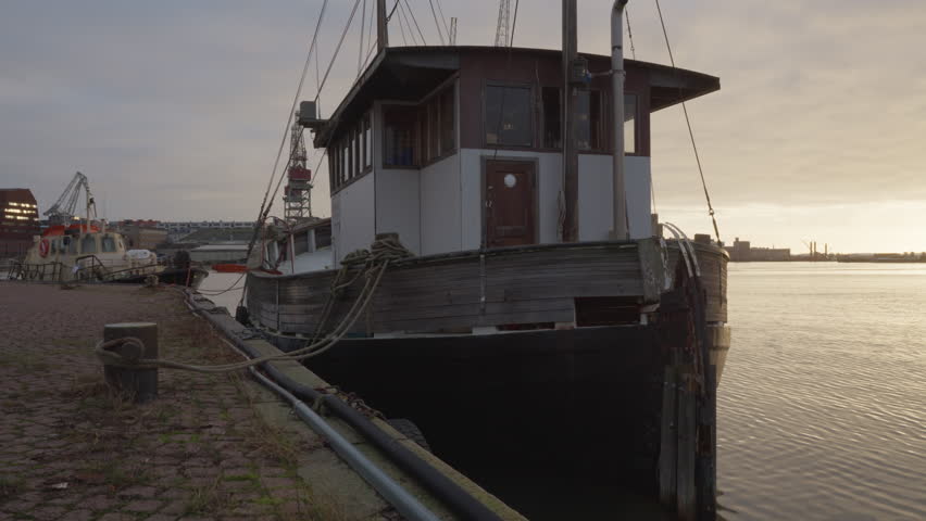 Old wooden boat moored at stone pier in Helsinki Ruoholahti harbour at sunset, vintage vessel docked in city port with industrial background under autumn overcast sky in Finland.