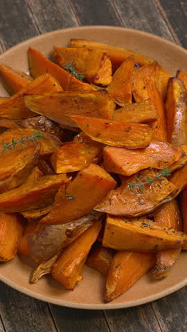 Roasted unpeeled sweet potatoes seasoned with thyme on a plate. Healthy vegan meal. Rustic wooden background, table spin. Vertical video.