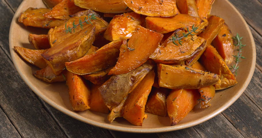 Roasted unpeeled sweet potatoes seasoned with thyme on a platter. Healthy vegan meal. Rustic wooden background, table spin.