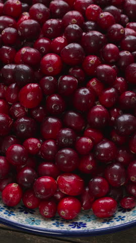 Close-up of fresh cranberries displayed on a decorative plate against a rustic wooden background, table spin. Vertical video.