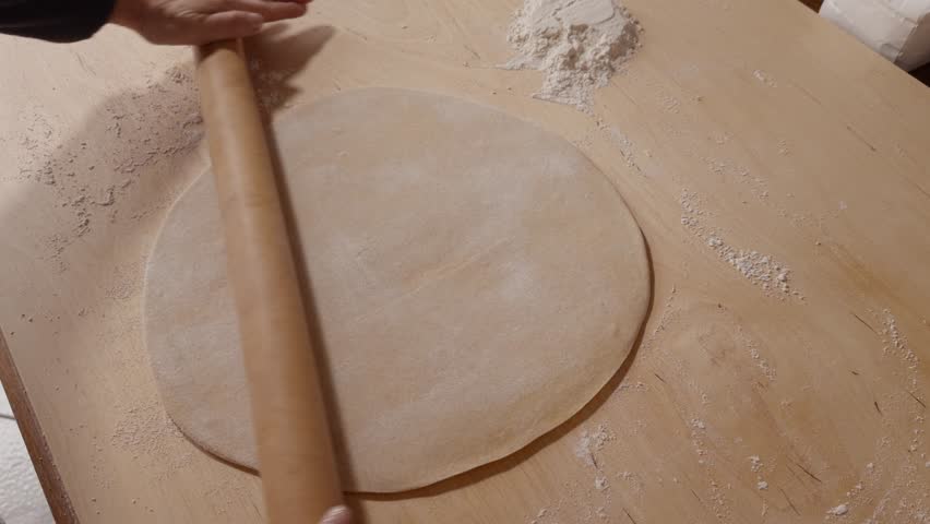 Senior hands using a wooden rolling pin to flatten dough on a floured surface, preparing homemade pasta or pastry.