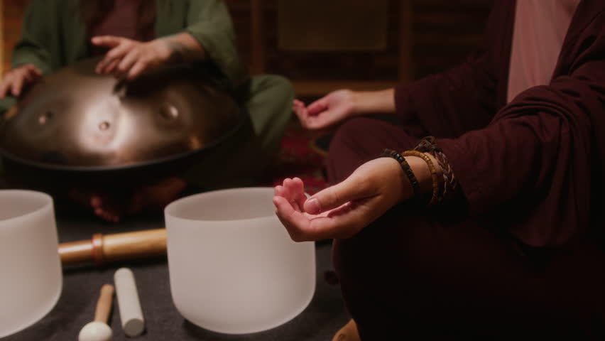 Medium shot of hands of unrecognizable young woman sitting with crossed legs in lotus posture on floor in studio, meditating during sound healing session, practitioner playing handpan in background