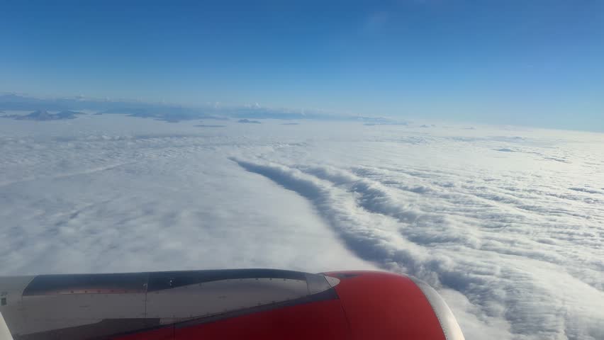 View of airplane engine wing flying above dense clouds with blue sky and distant mountain peaks. Aerial perspective of flight over cloud cover and mountainous landscape.