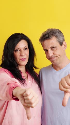 Mature man and woman showing disapproval with thumbs down gestures. Negative feedback and dissatisfaction concept on a bright yellow background. Vertical footage