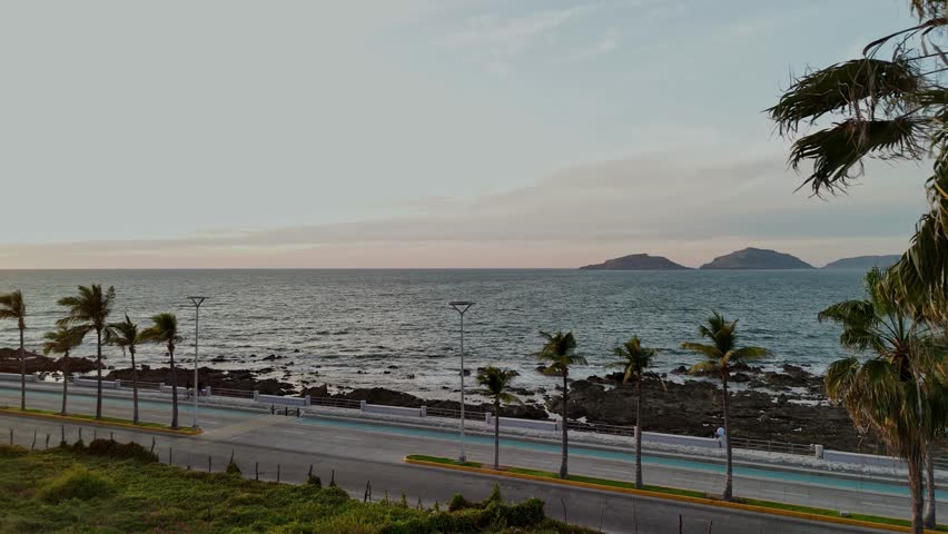 The sun approaches the horizon during sunset in Mazatlán, Sinaloa, casting warm golden light over the Pacific Ocean and the coastal boardwalk. The scene features oceanfront buildings, palm trees swaying in the wind, and sunlight reflecting on the water, creating a vibrant tropical atmosphere of a Mexican seaside city.