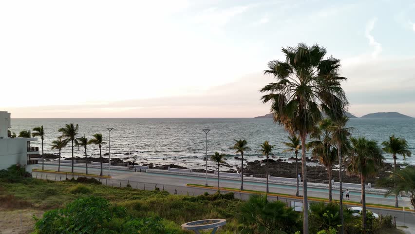 Panoramic view of the Pacific Ocean in Mazatlán, Sinaloa, at sunset. The scene shows calm sea waters, offshore islands, and the city skyline in the distance beneath a blue sky with clouds and airplane contrails lit by the warm evening light. A peaceful coastal landscape capturing the beauty of Mexico’s northwest coastline.
