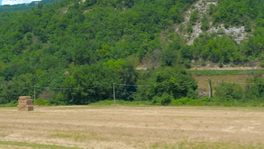 Hay bales standing in cultivated field near green hills. Rectangular hay bales stand drying in a harvested field, with lush green hills and trees in the background, creating a peaceful rural landscape