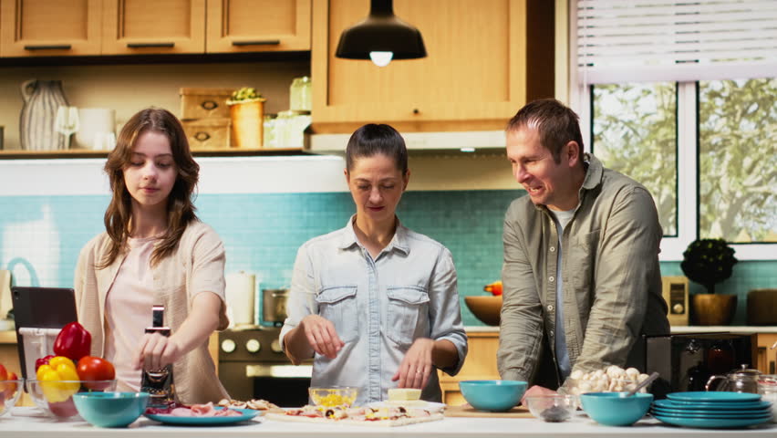 Tween daughter taking a family photo of her and parents during cooking time, smiling for a picture while they prepare homemade pizza in the kitchen. Fun weekend activity together. Camera B.