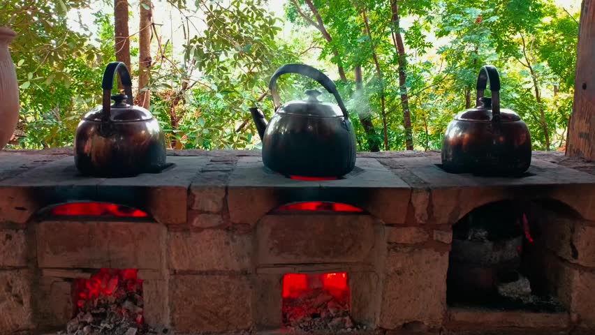 Static camera footage of three metal kettles boiling drinking water on a traditional wood fire stove in a rural village. Rustic daily cooking activity.