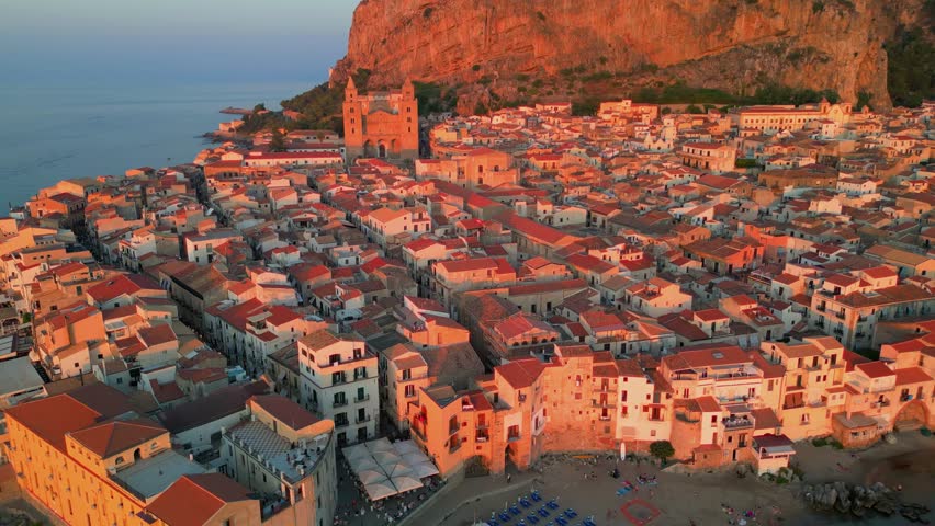 Aerial view of Cefalù revealing a dense old town, sandy beach, and calm turquoise waters along the Sicilian coast during the sunset