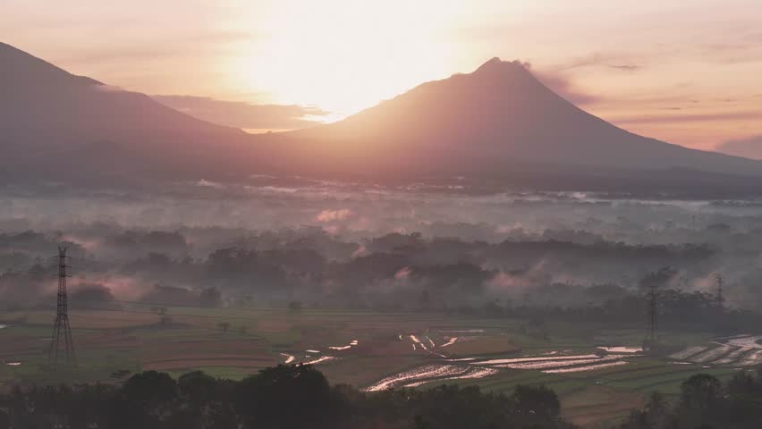 Drone footage of morning sunrise over mountains, foggy countryside, and reflective rice paddies in a peaceful rural scene.