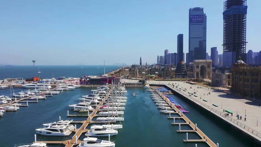 Aerial view shows a marina filled with sailboats near a waterfront promenade and the cityscape of Qingdao, China on a sunny day. People stroll along the walkway.