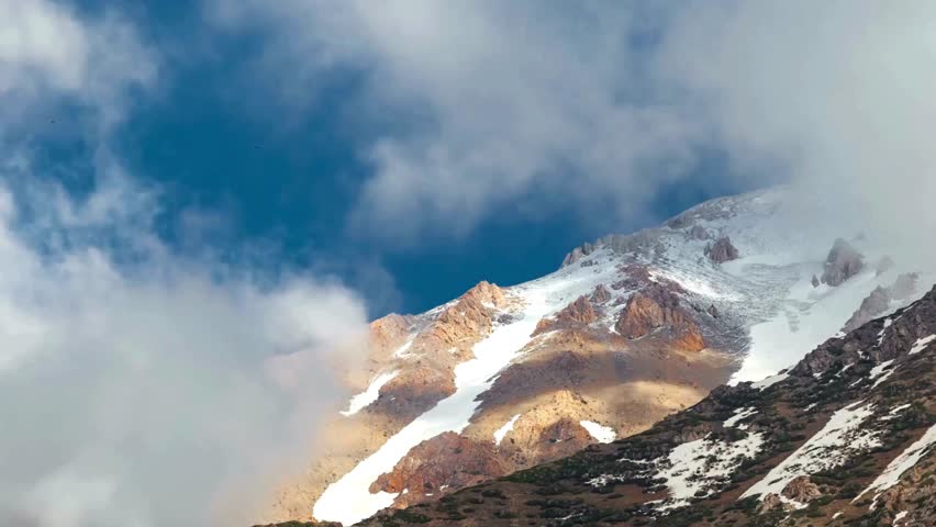 Breathtaking view of rugged mountain terrain featuring snow-capped peaks and dynamic clouds drifting across a clear blue sky. Perfect setting for nature lovers.