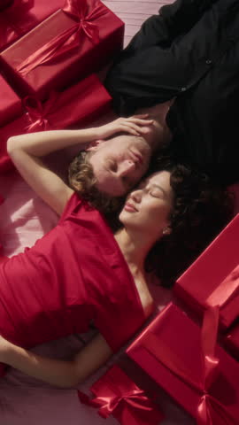 Vertical high angle view of elegantly dressed young lovers with their eyes closed lying on bed around present boxes, with girls hand on boyfriends neck