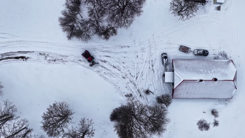 Top-down timelapse of a tractor clearing snow around a rural farmhouse, carving tracks across a white yard with parked cars and bare winter trees.