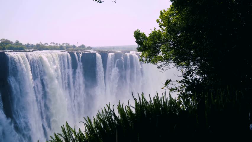 Water rushes dramatically over cliffs, creating mist in air amidst vibrant green foliage and blue skies. Serenity of nature captured in stunning detail.
