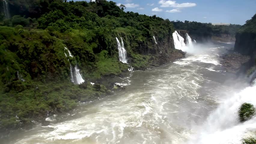 This captivating view showcases powerful waterfalls flowing into a river, framed by dense greenery and bright blue skies. The vibrant scene immerses you in nature