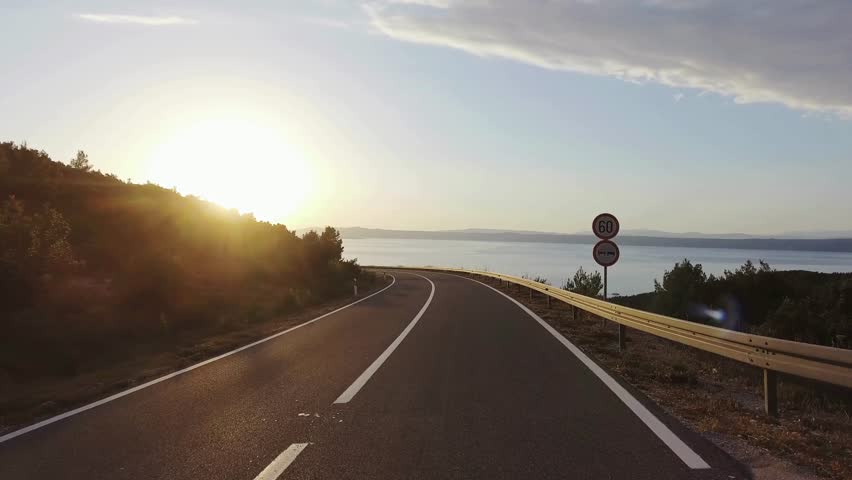 Camera moves along coastal road in Croatia during sunset. Beautiful Adriatic Sea views. Sun flares through the trees. Asphalt road curves along hillside.
