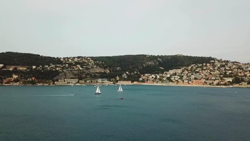 Several sailboats glide across blue Mediterranean waters off the French Riviera. Town extends up hillside, creating picturesque European coastal landscape.