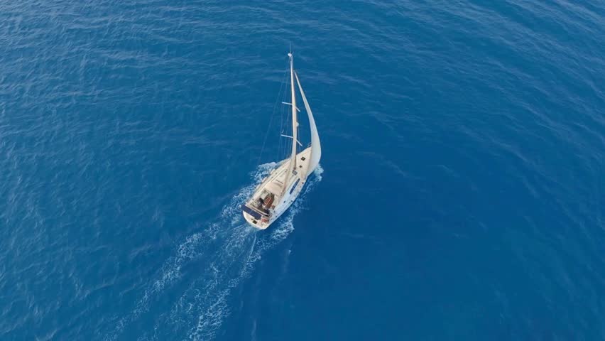 Sailboat with white sails is seen from above, moving across deep blue ocean. Sunlight reflects off the water's surface. Calm weather for sailing and enjoying the sea.
