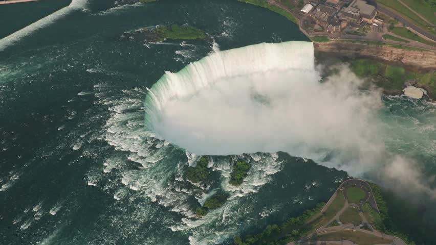 An aerial perspective captures the forceful rush of Niagara Falls as water plunges into the river. Greenery surrounds the falls, with a city visible in the distance.