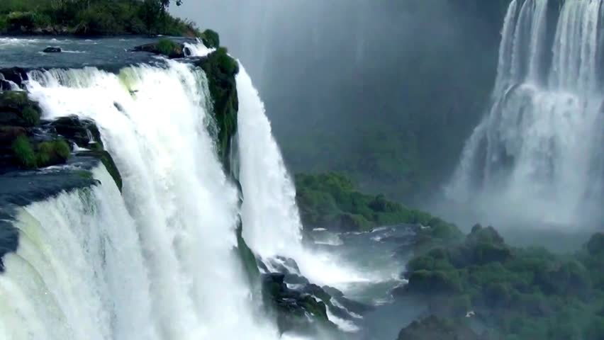 A daytime aerial perspective of Iguazu Falls shows several waterfalls cascading over rocks, surrounded by green foliage. Mist rises into the air creating a hazy atmosphere.