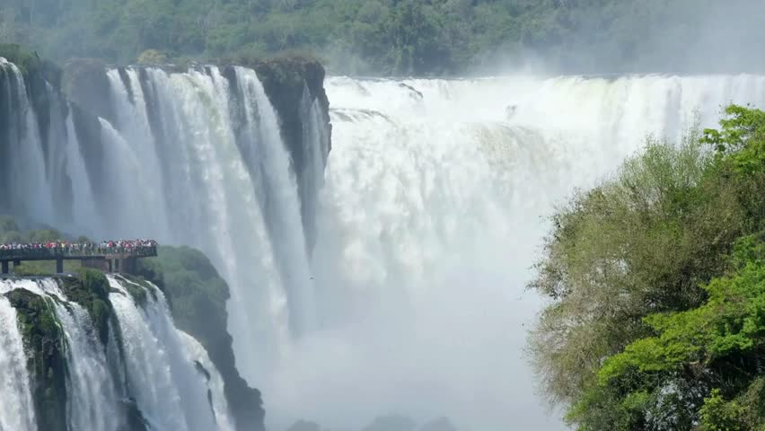 The sun shines on Iguazu Falls in Argentina. People walk along the observation deck, admiring the magnificent waterfalls and surrounding natural landscape.